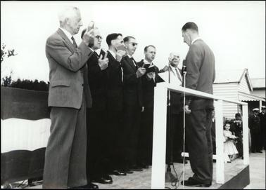 This image shows a group of six men standing before a seventh man at a swearing-in ceremony.  They are all holding a book in their right hand and the seventh man is reading from a sheet of paper.  Two microphones on stands are beside him.  Wooden buildings can be seen on the right-hand side of the photograph and people are standing in front of them, including a girl, holding a bouquet of flowers and two men in uniform.  