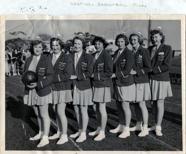 This image shows a group of seven girls in a basketball uniform posing for a photo.  They are all standing side-on with their left hand resting on the girl in front of them.  The first girl is holding a basketball with “Hastings” printed across it.  Their uniforms are light coloured short dresses with white collars, a blazer, a cap and white socks and sandshoes. The pocket on the blazer reads: “Hastings Basketball Club”.  People and motor cars are in the background.  The photograph has been glued onto a piece of white paper which has ‘Hastings Basketball Team’ printed across the top in pencil.  