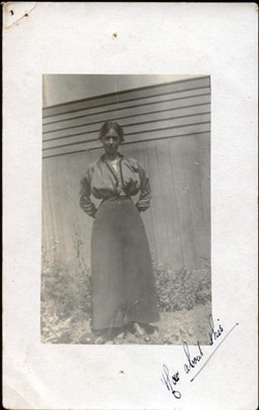 This image shows a woman standing in front of a high wooden construction, posing for a photograph.  She is wearing a long dark skirt, a long-sleeved jacket to the waist with a white blouse underneath.  Her hair is parted in the middle and pulled back.  Her hands are behind her back.  She is standing on grass and there is a flower bed behind her.  Written in ink across the bottom, right-hand corner is “how about this”.  
