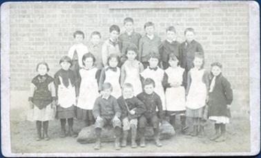 This faded image shows a group of children, in three rows, posing for a photograph.  There are seven children standing on a form in the back row, nine children standing in the middle row and three children sitting on a mattress in the front row.  There is a brick wall behind the children.  All the children are wearing coats or long sleeves and all are wearing boots.  The girls are wearing white aprons.  