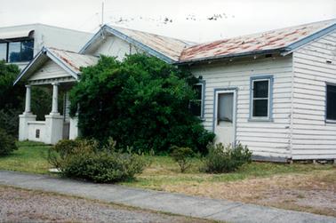 This image shows a weather board house taken slightly side-on.  A small verandah is at the front with a low concrete wall with two small columns. A small door with a single window on each side can be seen at the right-hand side of the building and part of a window is seen on the side of the house.  The roof is rusty.  A very large shrub is at the front against the house and small shrubs are on the front lawn.  A concrete footpath is in the foreground.  Part of a high house can be seen to the far left of the house.  A television antenna is on the roof.  