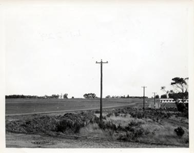 This image shows a large paddock pegged out with white pegs. There are trees in the background.  There is a road across the centre of the image with power poles along the right-hand side of the road.  A long white building can be seen halfway along the road on the right-hand side of the photograph and large trees are behind.  There appears to be a pile of dirt in the foreground on the left-hand side and grass and low scrub on the right-hand side.  A large gum tree is on the right-hand side of the image .  