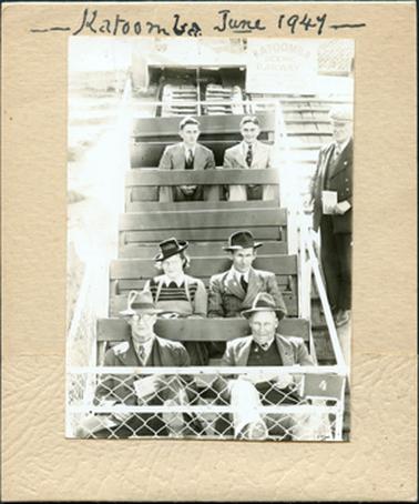 This image shows a group of six people sitting in an open railway carriage.  The two older men in the front seat are wearing hats and three-piece suits.  They are holding a piece of paper in their hands.  Their feet are on the wire cage in front of them.  The couple behind them are a woman and man, both wearing hats. He is wearing a suit and tie and she has a white collar over a pullover and is wearing spectacles.  The two younger men in the back seat are both wearing suits and ties.  An elderly man with spectacles and dressed in a three-piece suit and cap, holding a piece of paper in his left hand, is standing at the side looking on.  A faded sign above the carriage reads: “Katoomba Scenic Railway”.  Written in ink on the mounting board at the top is : “Katoomba June 1947”.