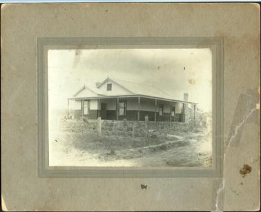 This image shows a house of timber construction on the bottom half of the walls and fibro cement on the top.  There is a large brick chimney on the right-hand side of the photograph and a tiny bit of chimney can be seen on the left of the house.  There is a vent near the roof at the front of the house.  There are timber verandah posts across the front and side of the house.  Boards fill in the area between the verandah and the ground.  Gardens can be seen to the front and side of the house.  There are some fence posts and wire netting across the front.  A roadway can be seen on the right-hand side foreground of the image.  