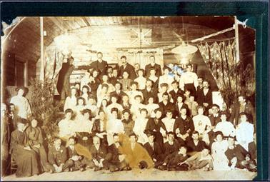 This image shows a very large group of people in front of a stage, inside a hall, posing for a photo.  There are approximately seventy people of all ages.  They are all very well dressed.  The back wall of the stage and a curved ceiling can be seen as well as drawn-back curtains on each side of the stage.  Two lanterns are handing above the stage and a pot plant is on either side of the stage.  