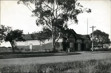 This image shows a side view of the Hastings Hall, taken from across the street.  The name is above the front door in large white printing.  The walls of the building have dark bricks half-way up.  There are three sets of windows along the side wall and a small door.  Two large sets of three windows are on either side of the front door.  A tall flag pole is attached at the front of the building.  A electricity pole is on the footpath.  There are two bench chairs on the footpath under trees on the left-hand side of the image.  Part of another building can be seen on the far right-hand side of the image.  Untidy grass is beside the road in the foreground.  