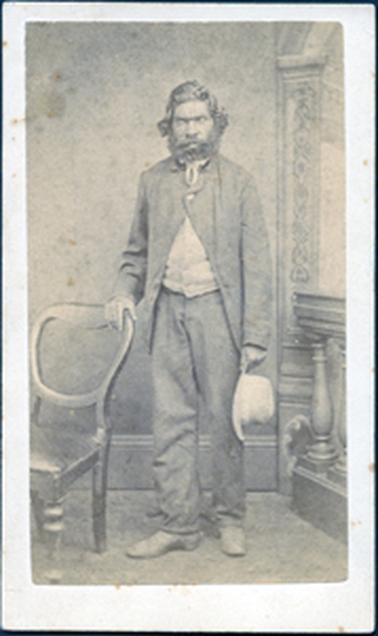 This image is of an aboriginal man posing for a studio photo.  He is standing beside a chair.  His right hand is resting on the back of the chair.  He is well dressed in long trousers, coat, leather shoes and buttoned vest with a tie at the neck.  He is holding a hat in his left hand.  His hair is wavy and he has a beard and moustache.  