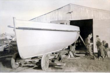 This image shows a boat on a trailer parked on grass in front of a large tin shed.  Two men and a small child are looking at something on the ground beside the boat and another man is walking away towards the shed. A man’s legs can be seen sticking out from under the trailer.  Part of a building can be seen on the far left-hand side of the image.  