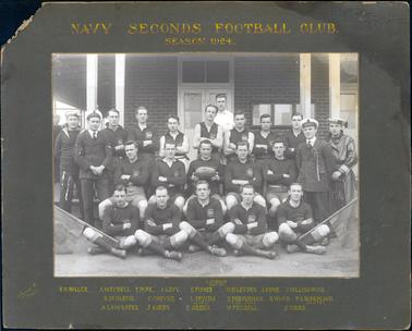 This image shows a group of footballers, in three rows, posing for a photograph.  There are twelve men in both players and navel uniform standing, five players sitting on chairs and five players sitting cross-legged on the ground.  All the players have their arms folded except for the player in the centre holding the football.  The football uniform is dark with the Australian Coat of Arms on the left-hand side of their shirt.  Two flags on short poles are being supported by two players in the front row.  The players are in front of a dark brick building which has three notice boards on the wall.  Two sets of doors and four white posts can also be seen.  The words “Navy Seconds Football Club. Season 1924.” is printed in gold letters on the mounting at the top and the names of the players and the photographers details are printed in gold on the mounting at the bottom.  
