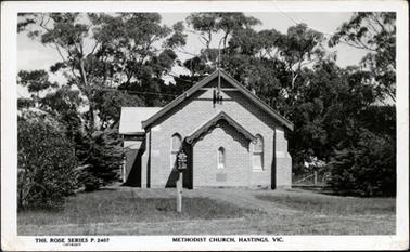 This image is of the Methodist Church in Hastings.  It shows a brick building with a notice board on the right-hand side of the front section of the Church.  There is grass all around with large shrubs on either side and large trees behind the church.  Part of a fence can be seen in the background.  Printed at the bottom of the postcard is: “The Rose Series P. 2407 Methodist Church, Hastings. Vic.”.