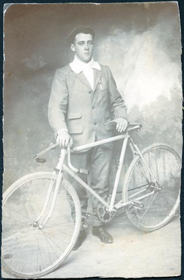 This image shows a young man, very smartly dressed, standing holding onto a bicycle with both hands.  He is wearing a suit and a white shirt with no tie.  The large high collar on his shirt is turned over his coat collar.  His coat has three buttons, two of which are done up.  There are four pockets on his coat, three of which have flaps.  He is wearing a cap with no brim and he has a bar-pin, with something attached, on the right-hand lapel of his coat.  His trousers come in at the ankles and he is wearing dark coloured lace-up shoes.  The handlebars on his bicycle are turned down.  