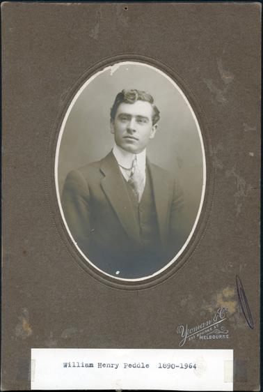 This is an oval portrait of a young man from the waist up.  He has wavy hair with a part on the left.  He is wearing a three-piece suit with a tie, which has a very small knot.  The collar on his shirt is very wide and rounded at the ends.  He is clean shaven and not looking directly into the camera.  The photographers name and address is printed in silver on the bottom right-hand side of the mounting.  A piece of white paper is taped to the bottom of the mounting with the typed words: “William Henry Peddle 1890-1964”. 