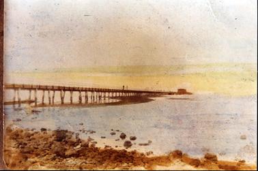 This image shows a jetty stretching out into the water, starting at the centre, left-hand side of the image and disappearing into the distance.  A building can be seen at the far end.  Two people can be seen in the distance on the jetty.  The tide is low as most of the structure is out of the water.  Large stones are in the foreground.  