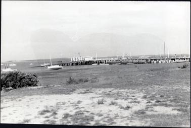This image is of a jetty, taken from the foreshore.  There are many boats beside the jetty as well as in the water in the foreground and on the left-hand side of the photograph.  A large boat can been seen behind the jetty in the distance.  There is a boat-shed on the end of the jetty.  A beach is in the foreground with mangroves on the left-hand side.  Land can be seen in the far distance.  