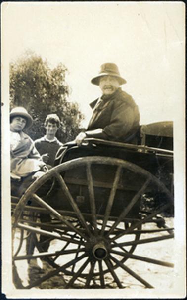 This image shows an elderly woman sitting in a phaeton, a young girl sitting opposite her and a young man standing behind.  The woman and girl are both wearing overcoats and hats.  Only part of the young man can be seen, but he is wearing a pullover.  They are all looking at the camera.  A tree can be seen in the background.  Only part of the vehicle can be seen.  