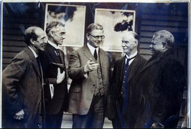 This image shows five men standing outside a weatherboard building.  Two sets of windows are behind the men.  The man in the middle of the group is talking and making a point with his outstretched right hand.  Everyone is smiling.  The three men at the centre are wearing three piece suits and the men at either end are wearing coats.  The three men on the left are wearing spectacles.  The man, second from the right, has a white moustache and the man on the far right has greying hair and dark coloured skin.  