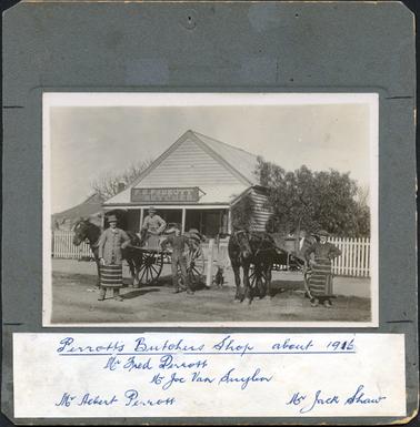 This image shows a weather-board  building with a galvanised iron roof and a verandah across the front with four posts.  A sign, F.G. Perrott Butcher, is on the roof of the verandah.  There is a picket fence on either side of the building.  A building can be seen on the left-hand side of the image and a bushy tree on the right-hand side of the image.  Standing posing for the photograph in front of the shop are four men.  There are two horses and carts. Two men are standing in front of the horse and cart on the left-hand side of the photograph and another man is sitting in the cart.  The man on the left is wearing a butcher’s apron, coat and hat and has his right thumb heavily bandaged.  The man in the cart is wearing a coat and hat and holding the reins and the man on the right is wearing high-waisted trousers with braces and a long sleeved shirt.  The man standing beside the horse and cart on the right-hand side of the photograph is wearing a butcher’s apron, coat, white shirt and tie and a cap.  They are all standing on the roadside and there is a footpath behind them.  There are two dogs in the photograph.  One is lying beside the fence and the other is sitting between the two carts.  Written in blue biro on white paper and glued to the bottom of the mounting is: “Perrott’s Butchers Shop about 1916 Mr Fred Perrott Mr Joe Van Suylen Mr Albert Perrott Mr Jack Shaw”.