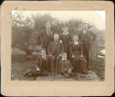 This image is a family portrait taken outdoors.  There are three people standing at the back, two people seated on chairs and two boys sitting on a rug on the ground.  Everyone is well dressed.  The two men at the back are wearing three-piece suits with ties and fob chains.  They each have one hand on their hip. The woman standing between them is wearing a long sleeved dress, the top having a wide collar that sweeps down to the waistline and has a lace collar at the neck.  The man sitting is wearing a three-piece suit with the coat buttoned at the top and a small black tie.  The woman beside him has and long-sleeved dress with a small capped sleeve, pleating on the bodice, a high white collar with lace bow.  The young man on the rug is wearing a three-piece suit as is the boy beside him who has a large white collar, small tie,  pants to the knee with leggings underneath.  Everyone has a stern look on their faces.  There are trees  behind them and some water can be seen on the right-hand side of the photograph.  