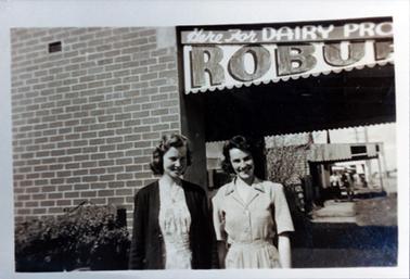 This image shows two young women standing beside a hedge, posing for a photo, in front of a shop.  The young woman on the left is wearing a print dress with a dark coloured cardigan.  The young woman on the right is wearing a short sleeved, checked dress with a revere collar.  Both women have wavy hair and are smiling.  The sign on the shop above their heads reads: Here for Dairy Pro Robur”.  The  brick wall of the shop is on the left-hand side.  Other shops and people on the footpath can be seen on the right-hand side.  The street is on the far right-hand side.  