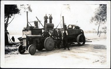 This image shows road-making equipment parked on the side of a gravel road.  A tractor is in the foreground, probably a Fordson.  A large trailer with drums, probably containing tar, is behind.  The trailer has large steel wheels.  Bitumen heating equipment is at the rear.  Two men are standing on equipment at the rear and in the foreground, one man is standing on the road and another sitting beside him on the trailer.  There is part of a tree on the left-hand side of the image and an item of clothing is hanging on a stick. There are trees on the right-hand side of the image.  