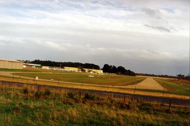 This image shows an airfield with a runway on the right-hand side.  Several buildings and small aircraft are on the left-hand side of the image.  Tall trees are in the background.  A bitumen road with a post and wire fence on either side is in the foreground beside a grassed paddock.  