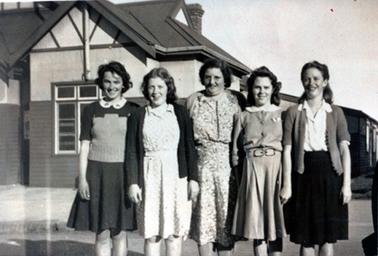 This image shows five women standing outside a building posing for a photo.  They are all smiling for the camera.  An older woman wearing spectacles and a print dress, is in the centre.  The other four women are very young.  The girl on left is wearing a dark coloured skirt and a two tone pullover with short sleeves.  The next girl is wearing a print dress and a dark coloured cardigan.  The next young woman is wearing a plain, light coloured dress with a white collar and the girl on the far right is wearing a black skirt, white blouse and a cardigan.  