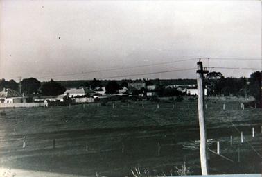 This image shows buildings in a town taken from a distance.  There is a telegraph pole with wires in the foreground and a large fenced paddock.  Houses, buildings, fences and trees can be seen.  The image is dark.
