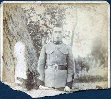 This photograph has been torn in half and only the top half remains.  The image is of a man in army uniform standing in front of a large tree.  His jacket has a high dark collar and dark cuffs on the sleeves.  It has two large top pockets.  He is wearing a wide belt with a large metal buckle.  There are trees and shrubs in the background.  There is a piece worn away on the large tree trunk on the left-hand side of the image.  