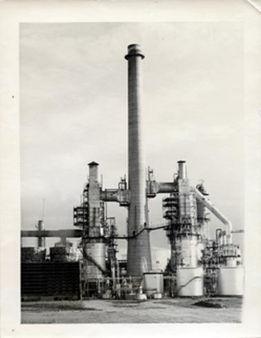 This image shows part of a refinery.  It has a very tall stack in the centre which is attached to two smaller structures on either side.  Other sections of the refinery are on either side and parts of large tanks can be seen in the background.  