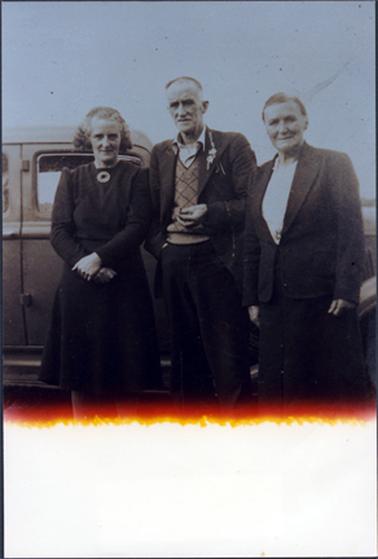 This image shows two women and a man posing for a photograph in front of a motor vehicle.  The woman on the left-hand side is wearing a long sleeved dark coloured dress with a large brooch at the neck.  She has her hands crossed at the front.  The woman on the right-hand side is wearing a dark suit with a white blouse.  Her hair is pulled back severely.  The man is wearing a suit with an open-necked shirt with a sweater underneath. He has a sprig of flowers in his coat lapel.  Light has been let into the bottom section of the photograph.