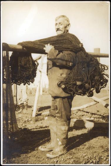 This image is of a fisherman, standing side-on, posing for a photo.  He is standing in front of a wooden rail with a large rolled-up fishing net across his left shoulder and hanging over the rail.  He is wearing a heavy duty coat and long trousers with knee-high rubber boots.  He has grey hair and moustache and a slight smile on his face.  Part of another fishing net can be seen on the far left-hand side of the image.  Part of a wire netting fence can be seen along with the legs of another man behind that.  