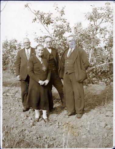 This image is of three men and one woman standing on rough ground in an orchard posing for the camera.  They are all dressed very well, the men in three-piece suits and ties and the woman in a long sleeved dress with a large collar and a large pleat in the front of the skirt.  She is wearing a brooch, white stockings and her hair is pulled back severely.  There are fruit trees behind them.  