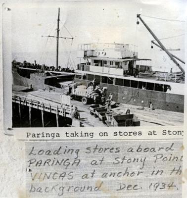 This image shows men loading bags of supplies from the back of a truck, parked on a jetty, onto a boat. Some men are standing around and a couple of men are sitting on the wharf watching.  There are several small wagons with supplies, parked on the wharf near the truck.  Some tall masts can be seen on both sides of the image. Four posts under the jetty can be seen on the left-hand foreground of the image.  Printed at the bottom of the image is: “Paringa taking on stores at Stony Loading stores aboard PARINGA at Stony Point VINCAS at anchor in the background Dec. 1934”.