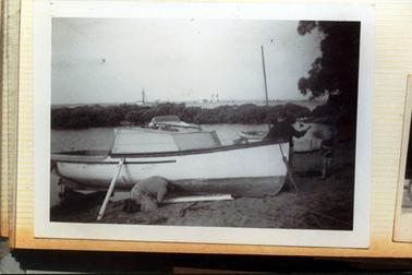 This image shows a wooden boat with a small cabin, standing on a sandy shore.  A man is kneeling beside the boat looking underneath.  Another man is standing at the front of the boat with his back to the camera.  His is holding out his right arm.  A small boy, on the far right-hand side of the image is walking towards the boat.  Part of another boat with a mast can be seen on the sand.  Water is behind the boats.  A line of trees is in the background and more water and some tall structures can be seen in the far background.  