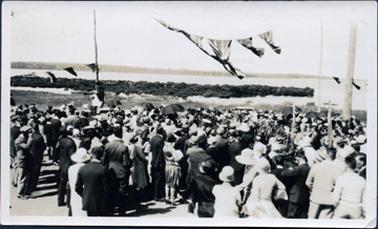 This image is of a very large gathering of men, woman and children with their backs to the camera.  All the people appear to be very well dressed and all are wearing hats.  Some umbrella’s can be seen.  There is a tall, thick post on the right-hand side with a thinner post behind it.  There are two lines of bunting attached to the posts and a line of flags is in the top centre of the image.  The foreshore is seen in the background.  