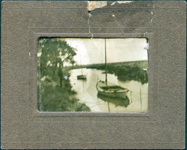 This is a blurred image of two boats in a creek.  There are trees on the left of the photograph where a man is seen standing.  There are low bushes on the opposite bank.  One boat is in the foreground with the second further down the creek.  Both boats have tall masts.  