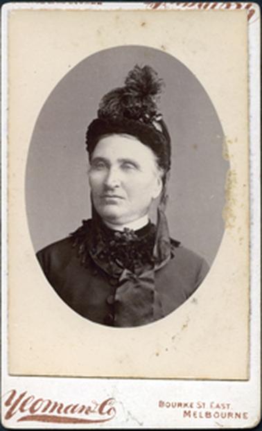 This image is of the head and shoulders of a woman wearing a high-necked garment with a tasselled collar and a wide satin tie. She has similar tassels on top of her bonnet.  She has a stern expression on her face. “Yeoman & Co Bourke St, East Melbourne” is printed at the bottom of the photograph.  