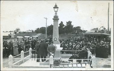 This image shows a tall war memorial with a lamp on the top standing in an enclosed area which is surrounded by eight posts with a chain attached.  Three men are standing on the left of the memorial and three men are sitting on the right-hand side.  They all have their backs to the camera.  A large crowd of people are standing and sitting facing the men.  A woman is sitting at an organ/piano on the right-hand side. Everyone is very well dressed and most are wearing hats and coats.  There are several vehicles on the left-hand side of the image and a bus with people looking out of the window on the right-hand side.  A street with houses to its left can be seen and more houses are partly visible on the right-hand side.  Tall trees are in the centre, background of the image.  Two telegraph poles can be seen.  