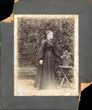 This image shows an elderly lady standing beside a small table. She has her hand on a pot plant which is in the centre of the table.  Her dress has long puffed sleeves and the skirt is touching the ground.  There are three rows of pleats around the bottom of the skirt.  A small lace collar is at the neck and the dress features satin ribbon let into the bodice and skirt.  Her hair is swept up at the back.  Part of a wooden building, covered in a creeper, can be seen behind her.  Wire netting is along the bottom of the building.  
