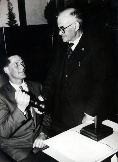This image shows two men dressed in suits and ties.  The younger man is sitting at a table and the older man is handing him a gavel.  They are both smiling.  The older man is wearing spectacles and has a badge pinned to his left lapel.  There is a small stand and papers on the table.  