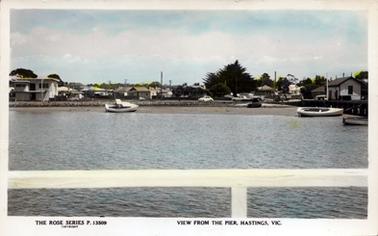 This image is a colourized postcard showing the view of a town from a pier.  The white railing of the pier is in the foreground.  There are some small boats in the water.  Houses, cars and trees can be seen in the town.  A fishing shed can be seen on the jetty on the far right-hand side of the image.  The sky is blue with some white puffy clouds.  Printed across the bottom of the postcard is: “The Rose Series P.13509 Copyright View from the pier, Hastings, Vic.”. 
