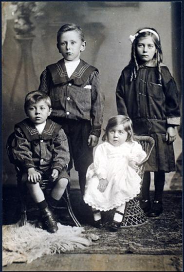 This image shows a portrait of four young children.  An older boy and girl are standing and a younger boy and girl are sitting on cane chairs in front of them.  The boys are dressed in sailor suits.  The eldest girl is wearing a dark long-sleeved dress with a light coloured pattern on the cuffs and the collar. She has long curls over her shoulders and a hair band with a bow on the side of her head.  The small girl is wearing a long-sleeved white dress with a lace trim on the bottom of her long sleeves, on the collar and around the hem of her dress.  All the children are wearing black shiny shoes.  There are two rugs on the wooden floor and a plant on a stand in the background.  None of the children are smiling.  