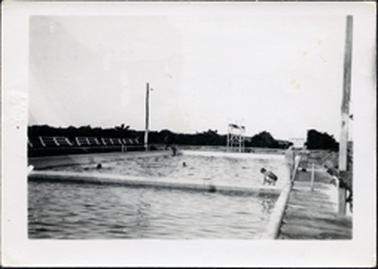 This very small image shows a swimming pool with a diving tower at the far end.  The pool is divided into two sections.  The heads of two people can be seen in the water.  A woman in bathers is crouching down beside the pool and two children are standing on the right-hand edge of the pool.  A walkway is along the right-hand side with a tall electricity pole in the foreground.  Another electricity pole with a light, is at the far left-hand side of the pool.  A line of fencing is on the left-hand side and trees are in the background.
