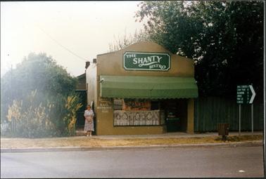 This image shows a small building with a woman standing beside it posing for a photograph.  The building has a large green sign at the top which reads: “The Shanty Bistro”.  There is a large green canvas blind across the front of the building.  There is a large window with a lace curtain in the lower half and a sign above it.  A front door is beside the window.  More writing is on the front of the shop to the left of the window.  The woman is wearing a print dress, white shoes and has her hands clasped at the front.  Part of a building can be seen at the rear of the shop.  A small tree with yellow flowers in front of it is on the left-hand side of the image. A large tree is behind a tall wooden fence on the right-hand side of the image. a brown rubbish bin and a road sign is on the footpath and a bitumen street is in the foreground. 