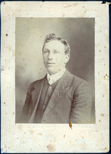 This image shows a young man posing for a photo, taken from the waist up.  He is turned slightly and not looking at the camera.  He is wearing a three piece pin-striped suit, white shirt and tie.  His fair hair is parted on the left-hand side and swept up on the right-hand side.  The photographers details are embossed in silver at the bottom of the mounting.