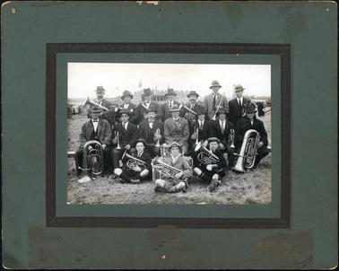This image shows a brass band posing for a photograph.  The group comprises of seventeen men and boys.  They are outdoors on the grass in three rows.  There are seven standing in the back row, seven sitting on a form in the middle row and three sitting on the grass in the front row.  They are all dressed in suits, white shirts and ties and hats.  Some are wearing coats and they are all holding their brass instruments.  Flags can be seen behind them beside a spectators stand.  Part of a building and water tank can be seen on the left-hand side of the image.  