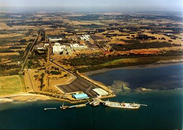 This aerial photograph shows an industrial plant on a foreshore.  The sea is in the foreground and a large ship is beside a wharf.  Sheds and smaller jetties are in the foreground and large sheds are in the middle of the image.  Large tanks can be seen on the left-hand side.  Trees, paddocks and housing are scattered across the top half of the image.  