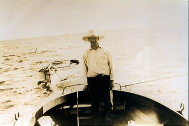 This image shows a man standing in the back of a wooden boat holding the rudder.  He is towing a small boat behind him.  The man is wearing a wide brimmed light coloured hat, long sleeved white shirt and dark long trousers.  He has a smile on his face and is looking at the camera.  Choppy water can be seen in the background.  