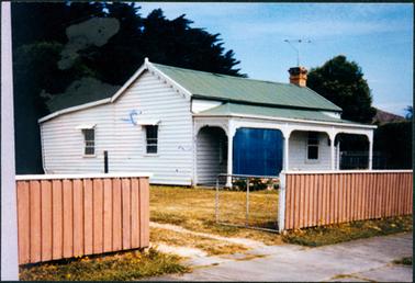 This image shows a small white cottage with a green roof.  A four post verandah is at the front with an arched lace trim from post to post.  There is a brick chimney on a top corner of the roof with a television antenna attached.  Two windows can be seen on the side of the cottage and two at the front.  A blind is pulled down on the left-hand side of the verandah.  Flowers can be seen in front of the verandah and tall trees behind the cottage.  A brown timber fence with a white rail on top is across the front.  A wire gate is open showing two tracks for the driveway.  A concrete footpath is in the foreground.  