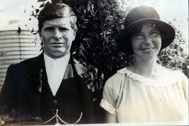 This image is a portrait of a man and woman taken from the waist up.  The man is wearing a white shirt buttoned at the neck, a waistcoat and coat.  A fob chain is attached to the waistcoat.  The woman is wearing a short sleeved dress with a round neck.  Lace is around the neck and sleeves.  She is wearing a dark coloured hat.  She has a smile on her face.  A galvanized iron rainwater tank and shrubs are in the background.  